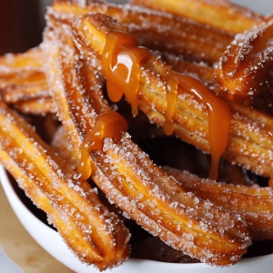 A stack of golden brown Salted Caramel Churros on a plate. They have salt sprinkled on them and caramel sauce dripped over them