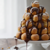 Golden profiteroles tower on a cake stand with chocolate sauce and powdered sugar
